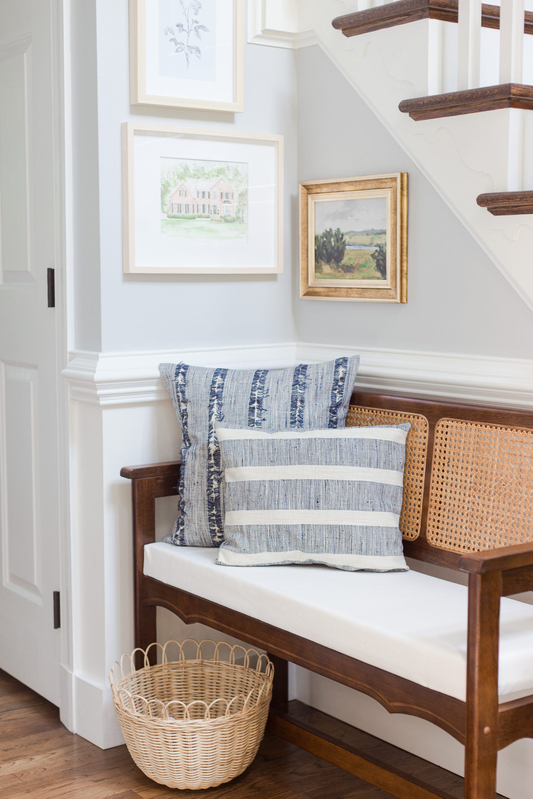 Two blue and white stripe pillows sitting on a cane bench in an entryway with pictures hung above the bench. 