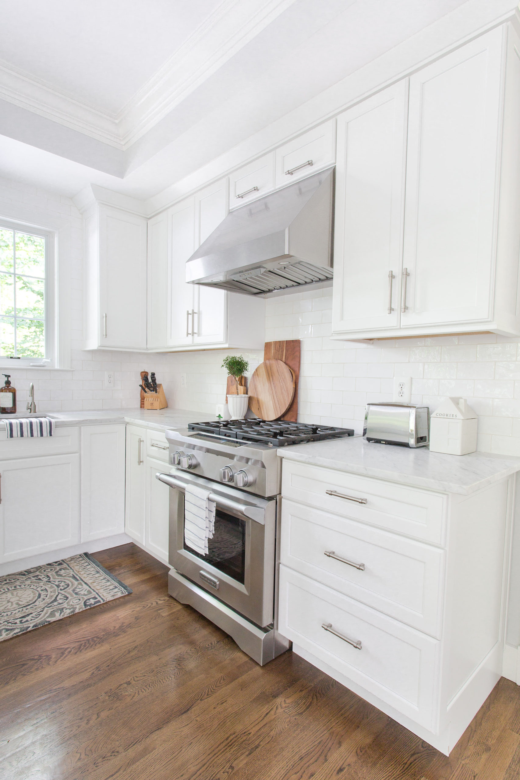 Gas range in white kitchen with dark wood floors. On the counter there is a cookie jar, wooden trays and toaster oven