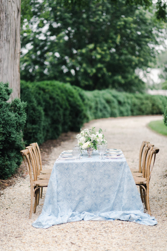 table set with blue tablecloth outside on pebble driveway with boxwoods surrounding. Wooden chairs and a floral centerpiece